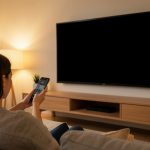 A person on a sofa holding a smartphone pointed at a wall-mounted TV showing a blank black screen in a warmly lit living room