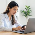 A person signing in on a laptop while holding a new smartphone at a home-office desk