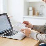 A person at a kitchen table checking coverage on a laptop while holding a mail-in health screening document
