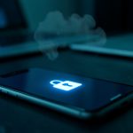A smartphone glowing with a padlock icon on a dark desk, a faint cloud shape above it and a blurred laptop behind