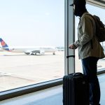 A traveler with a backpack and rolling suitcase standing at an airport gate window looking out at a plane on the tarmac