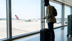 A traveler with a backpack and rolling suitcase standing at an airport gate window looking out at a plane on the tarmac