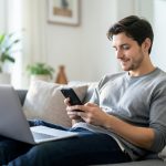 A person on a sofa checking their smartphone with a laptop open beside them