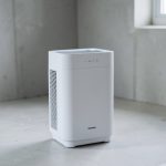 A white portable dehumidifier standing on the concrete floor of a bare basement with a small window in the background