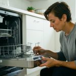 A homeowner kneeling at an open stainless steel dishwasher, checking the lower rack and filter area