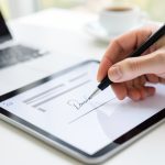 A hand signing a signature on a tablet screen with a stylus, a laptop and coffee blurred behind on a bright desk