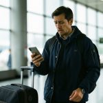 A traveler in an airport terminal looking concerned at a smartphone with a suitcase beside them