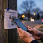 Hands holding a smartphone at dusk near a blurred missing-pet flyer on a pole