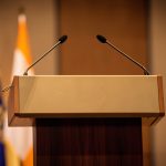 An empty conference lectern with microphones and blurred flags under warm lighting