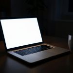 A laptop glowing in a dark home office at night beside a coffee mug