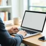 A person logging into a laptop with a blank screen while a smartphone rests on the desk beside it