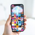A hand holding a smartphone showing a grid of photo thumbnails with a glowing cloud and upward arrow above the screen