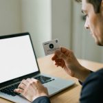 A person holding an ID card up to a laptop webcam to verify their identity for an online credit application