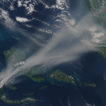 Aerial view of grey smoke plumes drifting from a green archipelago across the sea