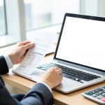 A person in business attire reviewing a paper invoice beside a laptop with a blank screen and a calculator on the desk