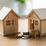 Two small wooden model houses beside a stack of coins on a wooden desk