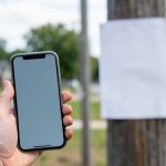A hand holding a phone with an incoming call beside a blurred flyer on a pole in a suburban street