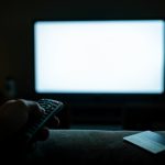 A hand holding a TV remote pointed at a glowing television in a dim living room with a credit card nearby