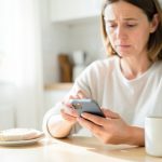 A person at a bright kitchen table tapping a smartphone with a puzzled expression, a coffee mug beside them