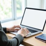 A person booking a hotel on a laptop with a blank screen, a passport and notebook on the desk beside them