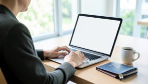 A person booking a hotel on a laptop with a blank screen, a passport and notebook on the desk beside them
