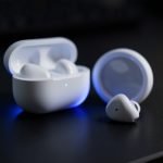 A pair of white true wireless earbuds with an open charging case on a dark desk, one earbud sitting apart
