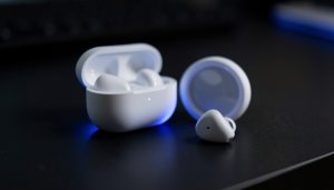 A pair of white true wireless earbuds with an open charging case on a dark desk, one earbud sitting apart
