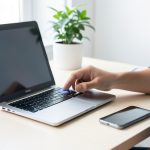 A person signing in to a laptop with a fingerprint sensor, a smartphone resting beside it on a bright desk