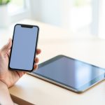 A person holding a smartphone with a blank screen while a tablet lies on the desk beside a coffee mug