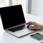 A person signing in on a laptop with a blank screen and a smartphone lying beside it on a clean desk