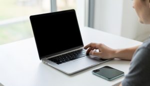 A person signing in on a laptop with a blank screen and a smartphone lying beside it on a clean desk