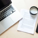 An overhead desk flat lay with a laptop, a stack of printed pages, a magnifying glass over a document, and a smartphone