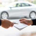 A car key and vehicle documents changing hands across a table with a sedan visible through the window