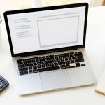 A bright freelancer desk seen from above with a laptop, calculator, notepad, and coffee