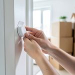 A person peeling the backing off a small wireless security sensor to stick on a door frame, with moving boxes behind