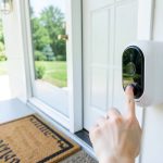 A white video doorbell camera mounted beside a residential front door with a visitor reaching for the button