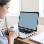 A person at a desk holding a coffee mug and looking at a laptop with a blank screen, a paper planner open beside it
