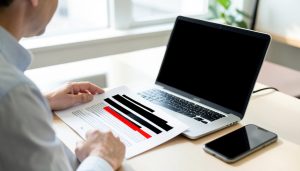A person reviewing a printed document covered with black redaction bars beside a laptop with a blank screen and a phone