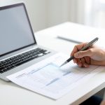 A person reviewing a printed spreadsheet of tabular data with a pen, beside an open laptop with a blank screen