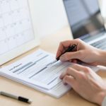 Hands filling out leave paperwork forms on a desk beside a wall calendar and a laptop