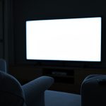 A blank glowing television screen in a dim living room with a remote on the sofa arm