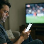 A person on a couch looking skeptically at a smartphone with a sports game on the television behind them