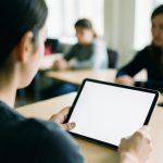 A student using a tablet at a classroom desk with classmates and a teacher blurred in the background