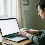 A person at a home desk looking at a laptop with a blank screen while waiting for a video appointment to load