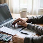 A person filing taxes at a home desk with a laptop, a printed tax form, a calculator, and a coffee mug