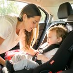 A parent leaning into a car's back seat to check a toddler buckled into a child car seat, with a suitcase visible nearby