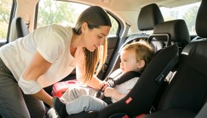 A parent leaning into a car's back seat to check a toddler buckled into a child car seat, with a suitcase visible nearby