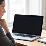 A person at a desk looking at a laptop with a blank screen and a puzzled expression, a coffee mug and notebook nearby