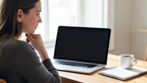 A person at a desk looking at a laptop with a blank screen and a puzzled expression, a coffee mug and notebook nearby