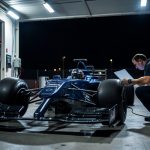 An open-wheel race car under bright inspection lights in a pit garage with a mechanic and laptop